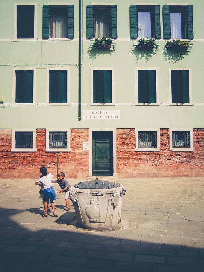Children playing beside a stone well in a sunlit square of pastel buildings